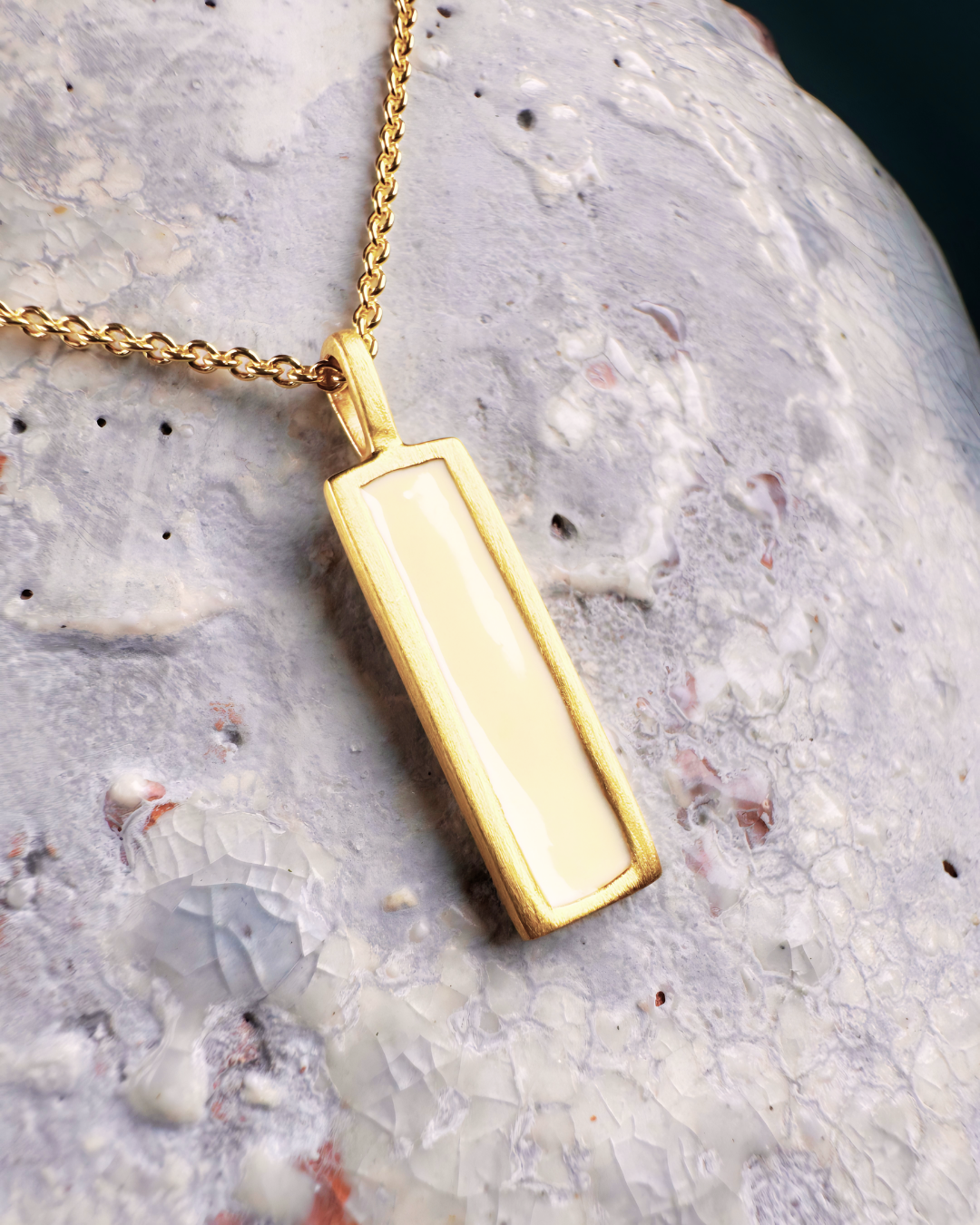 Gold-plated pendant with rectangular white enamel, displayed on a stone surface