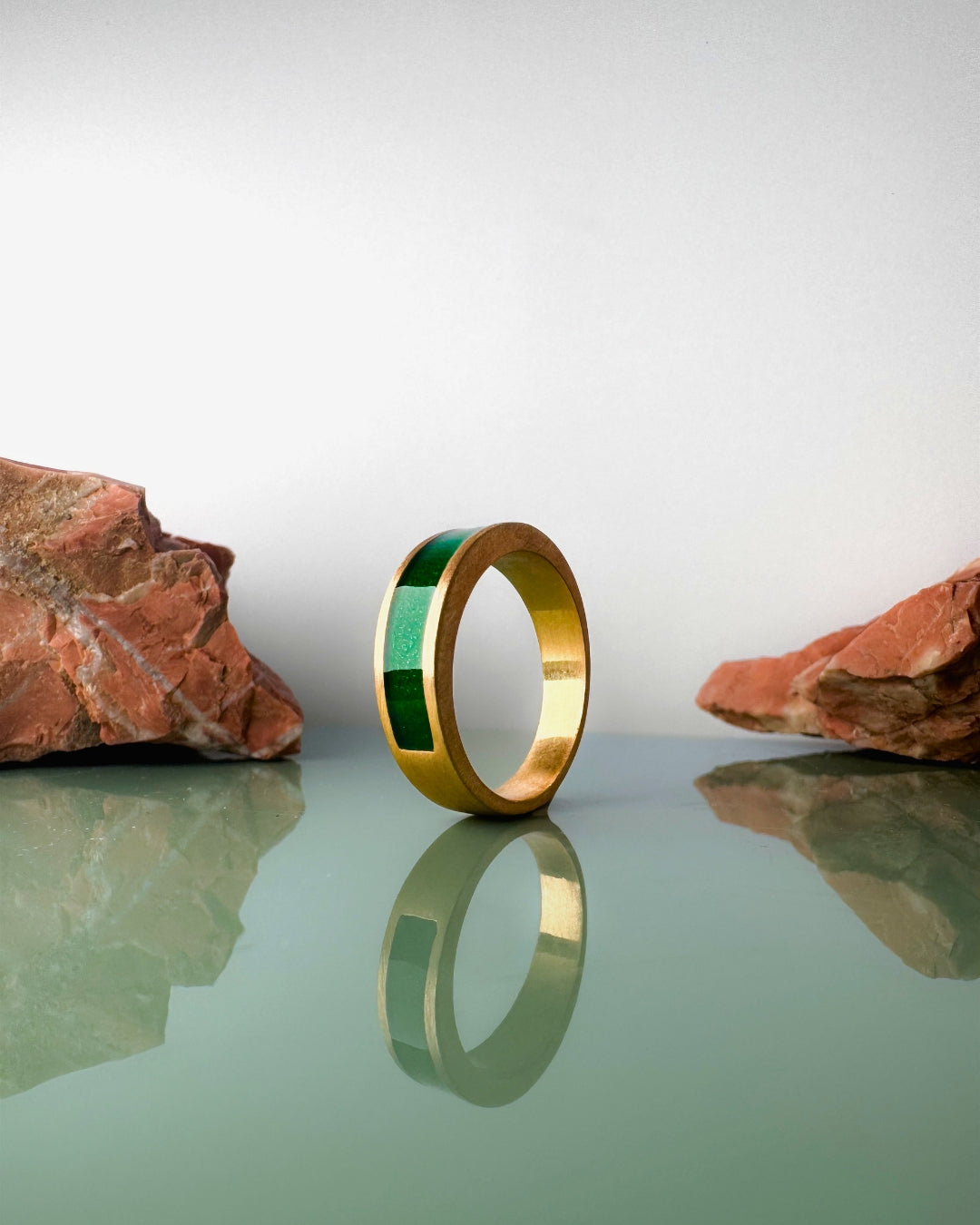 Gold-plated ring with a green enamel band on a reflective surface with rocks in the background